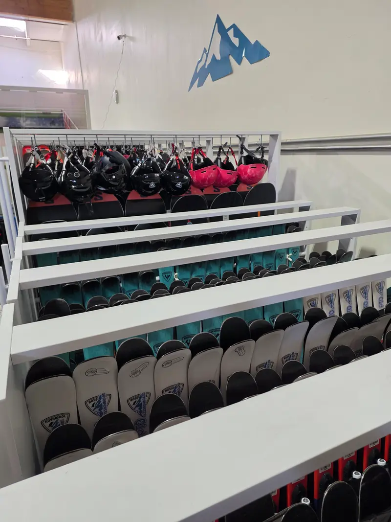 Rows of skis, including PTO-branded skis, with snowboards and hanging helmets beneath a blue mountain logo on the wall